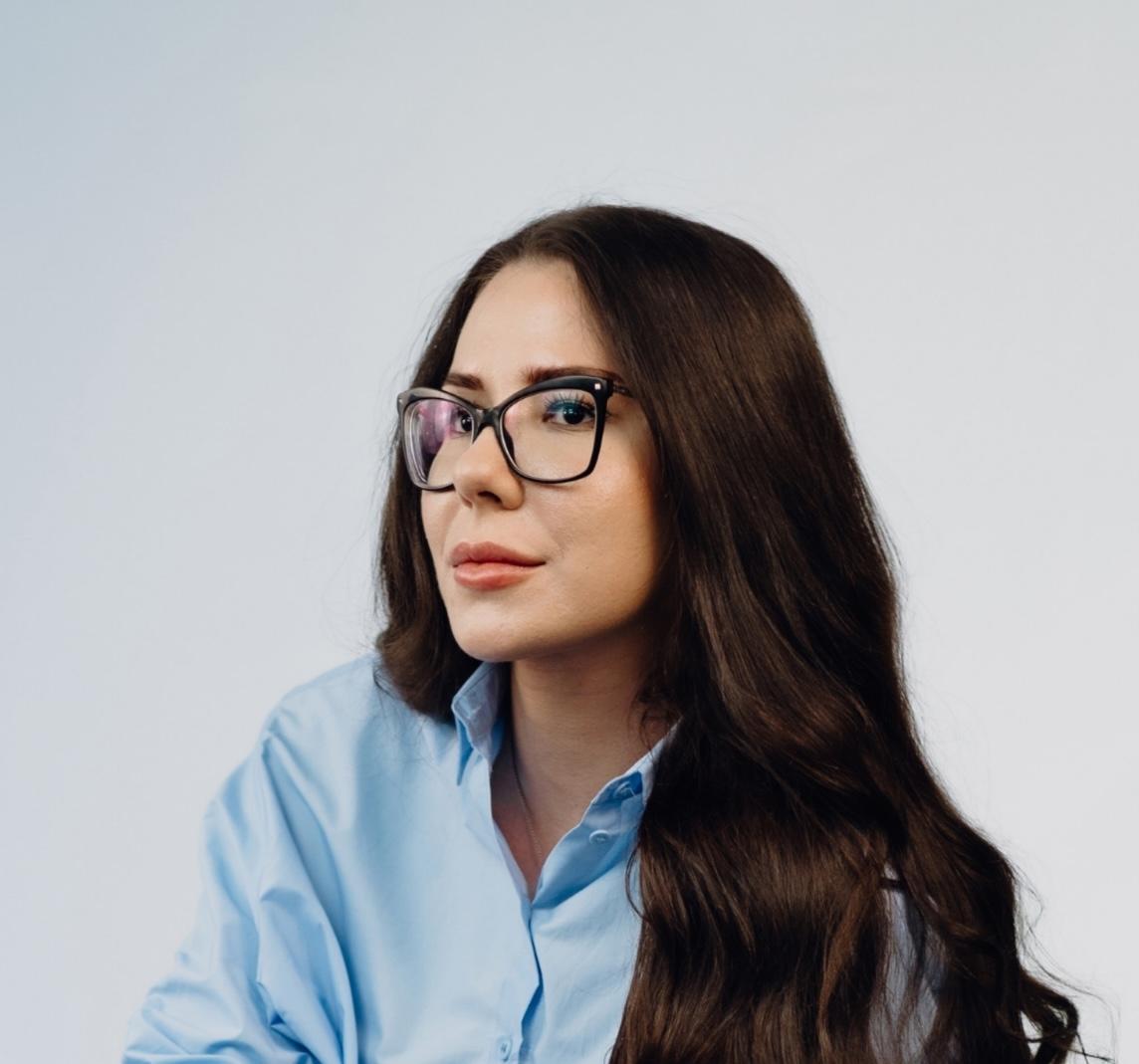 Professionelles Porträtfoto einer Frau mit langen dunklen gewellten Haaren und Brille, in einer hellblauen Bluse und dunkler Jeans, mit verschränkten Armen und leichtem Lächeln in die Kamera blickend.